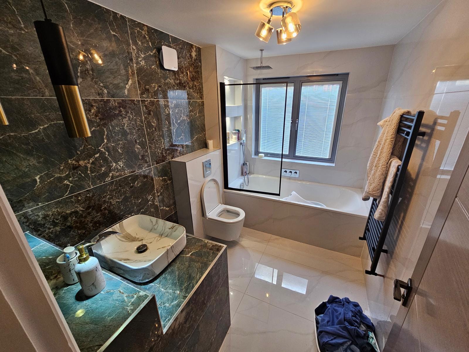 Bathroom with a bath and black-framed screen under a grey window, a vessel basin on a tiled plinth and a wall-hung WC.