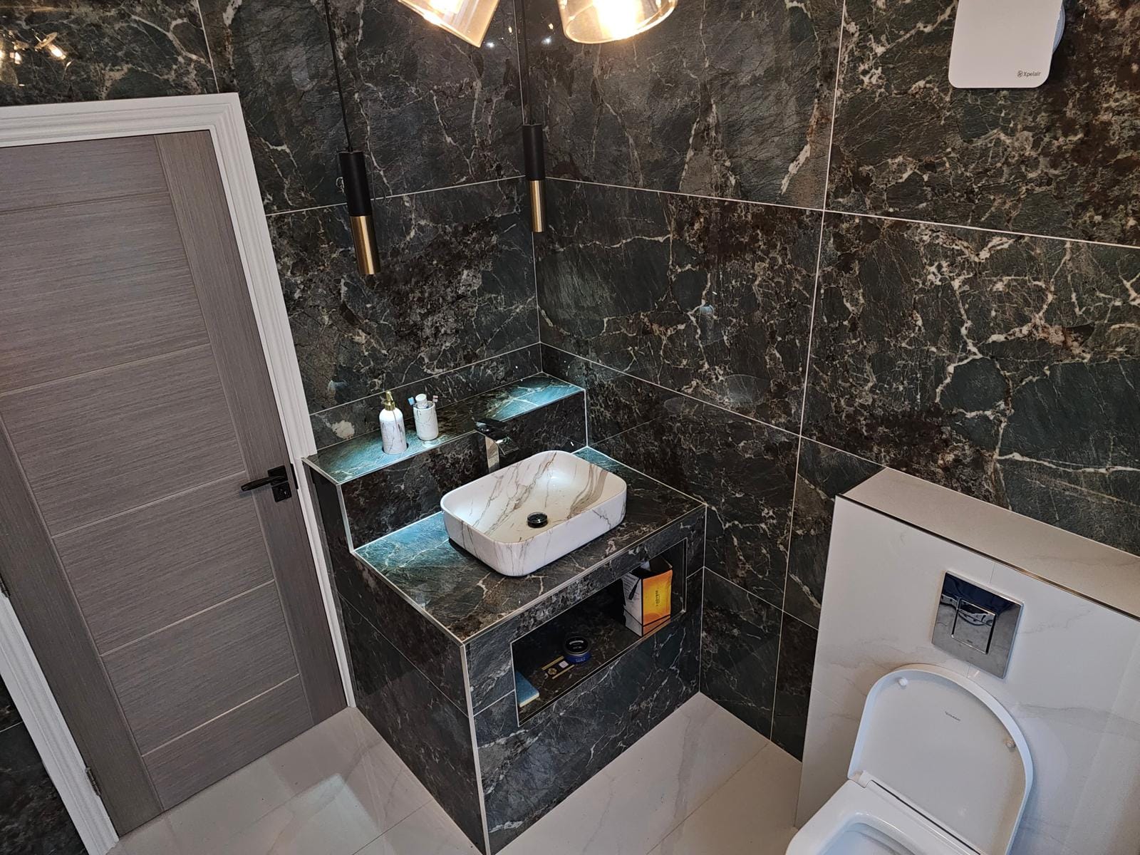 Vessel basin on a tiled plinth with a lit shelf and niche, beneath black-and-gold pendant lights.