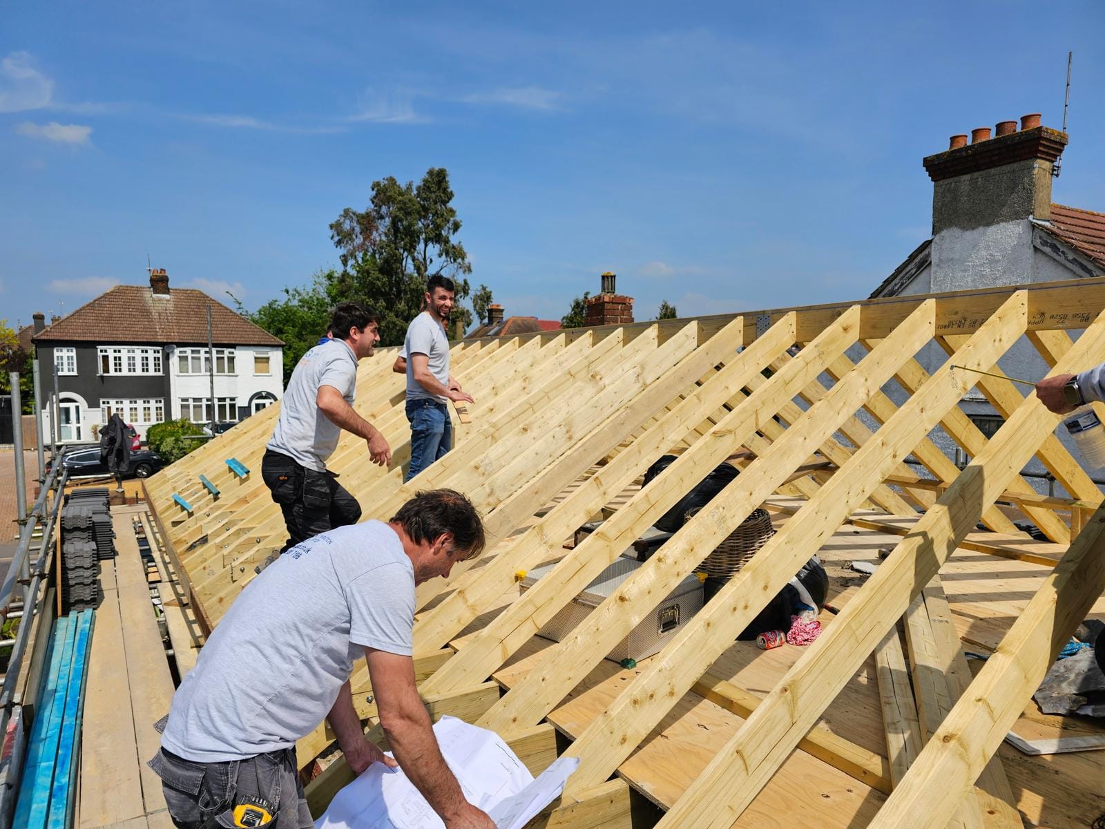 Carpenter leaning over roof drawings on the scaffold while two colleagues stand on the new rafters.