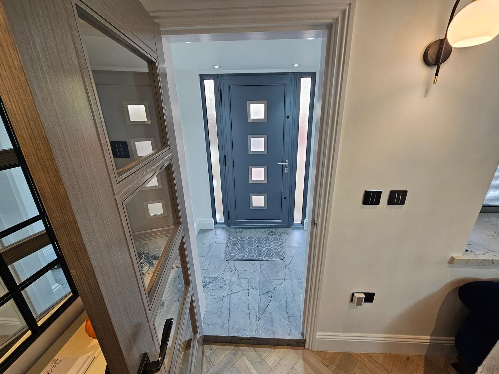 Entrance hall with a grey composite front door and glazed side panels, marble-effect floor tiles and herringbone flooring.