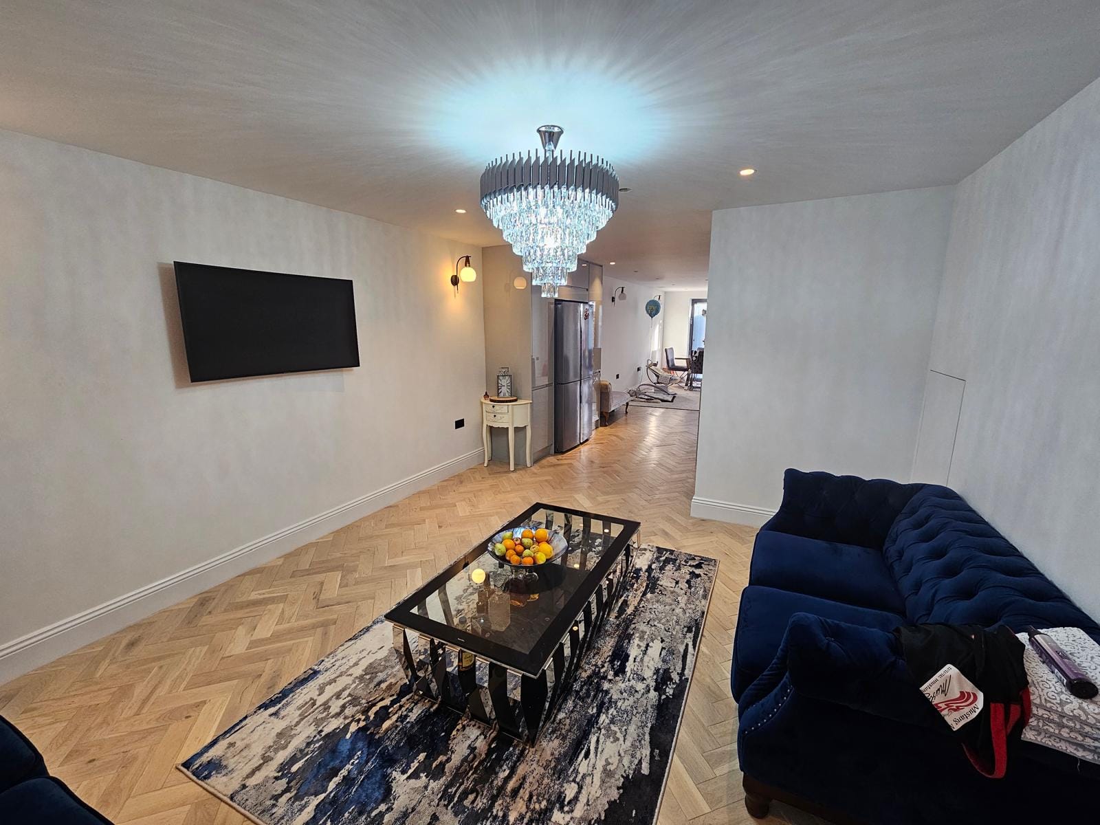 Living area with a tiered crystal chandelier, wall-mounted TV, glass coffee table and navy velvet sofa.