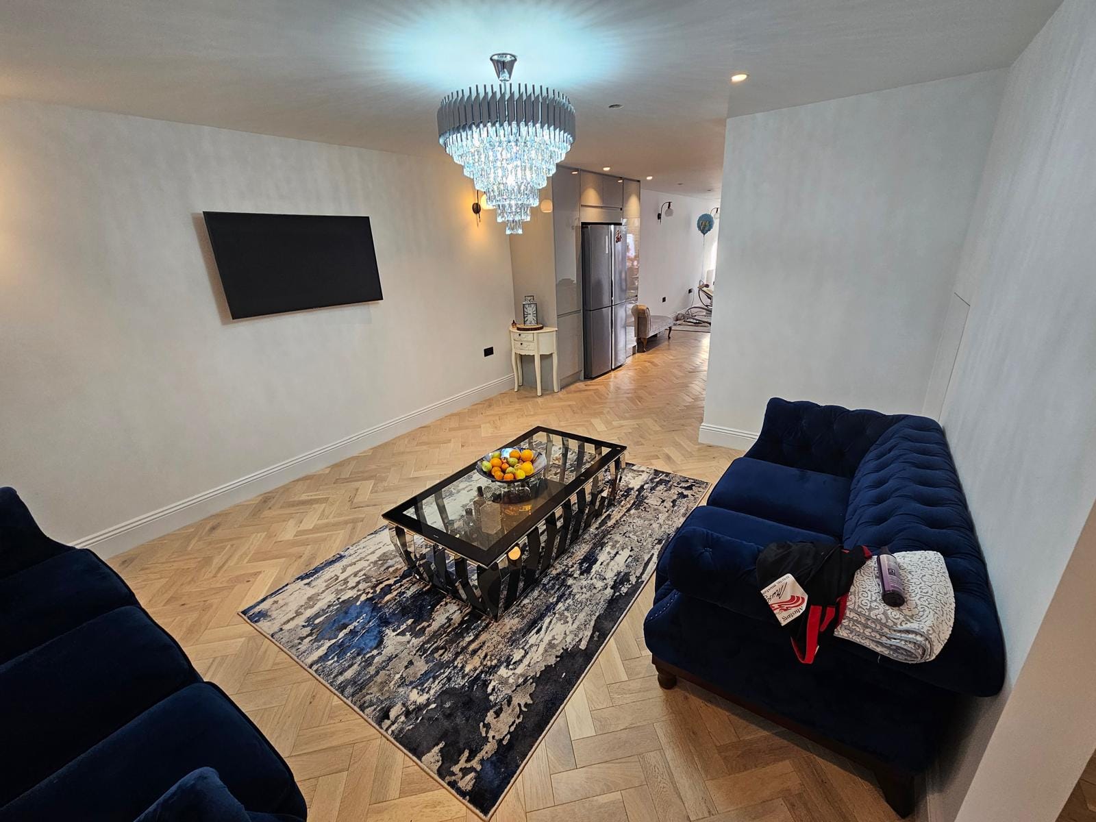 Open-plan living room with a crystal chandelier, navy velvet sofas, a glass coffee table and herringbone flooring.