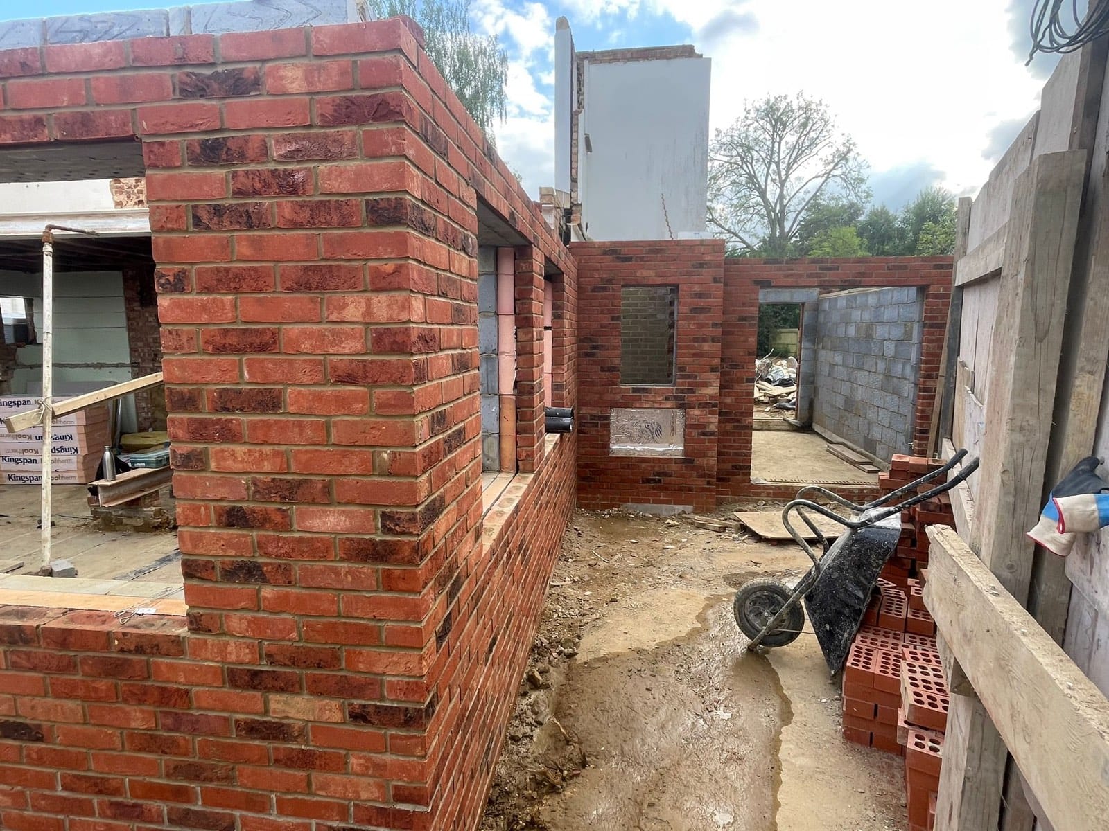 New-build brick-and-block walls with openings formed, bricks stacked and a wheelbarrow on site.