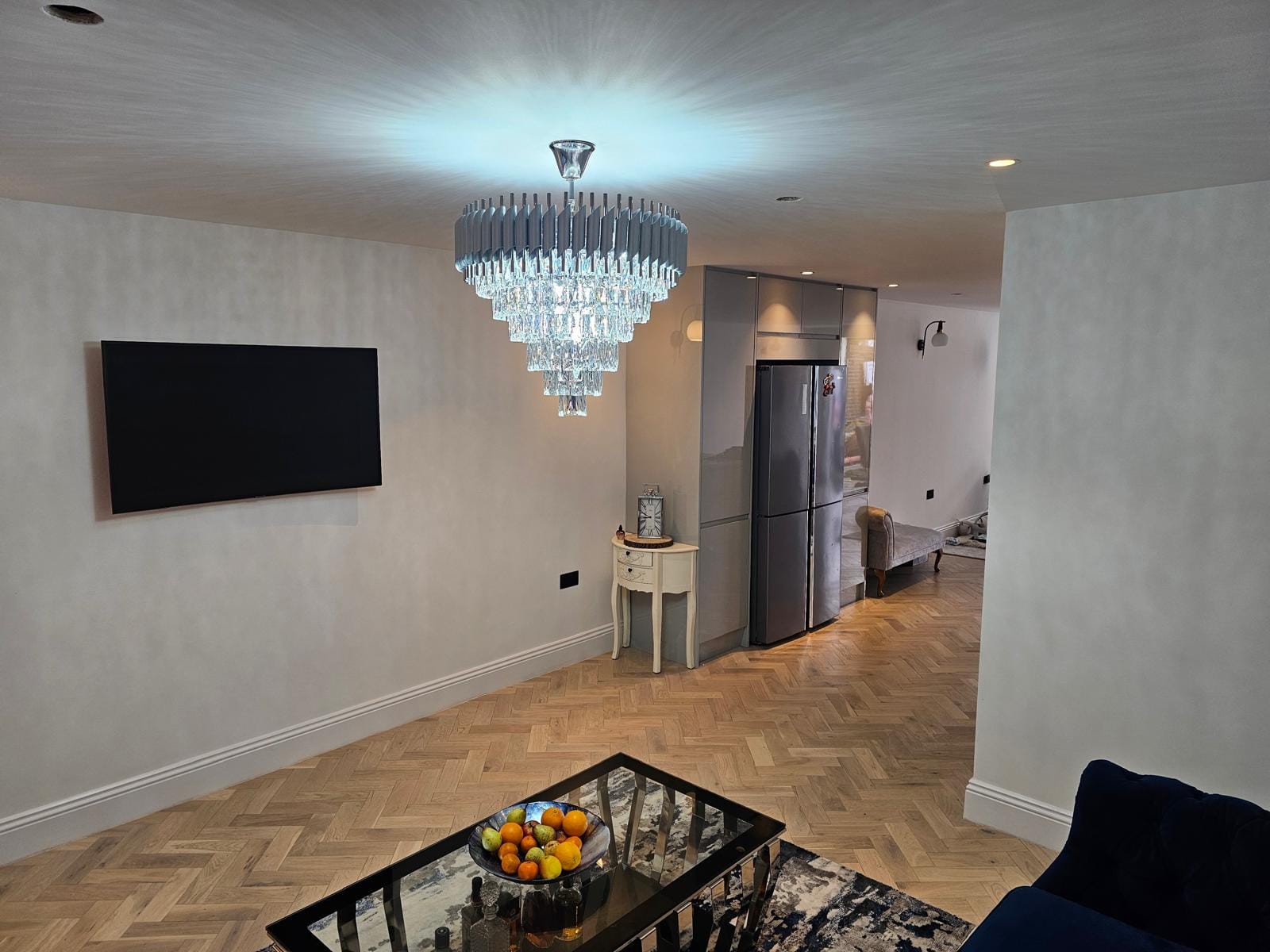 Open-plan living room with a tiered crystal chandelier, wall-mounted TV and herringbone floor leading to the kitchen.