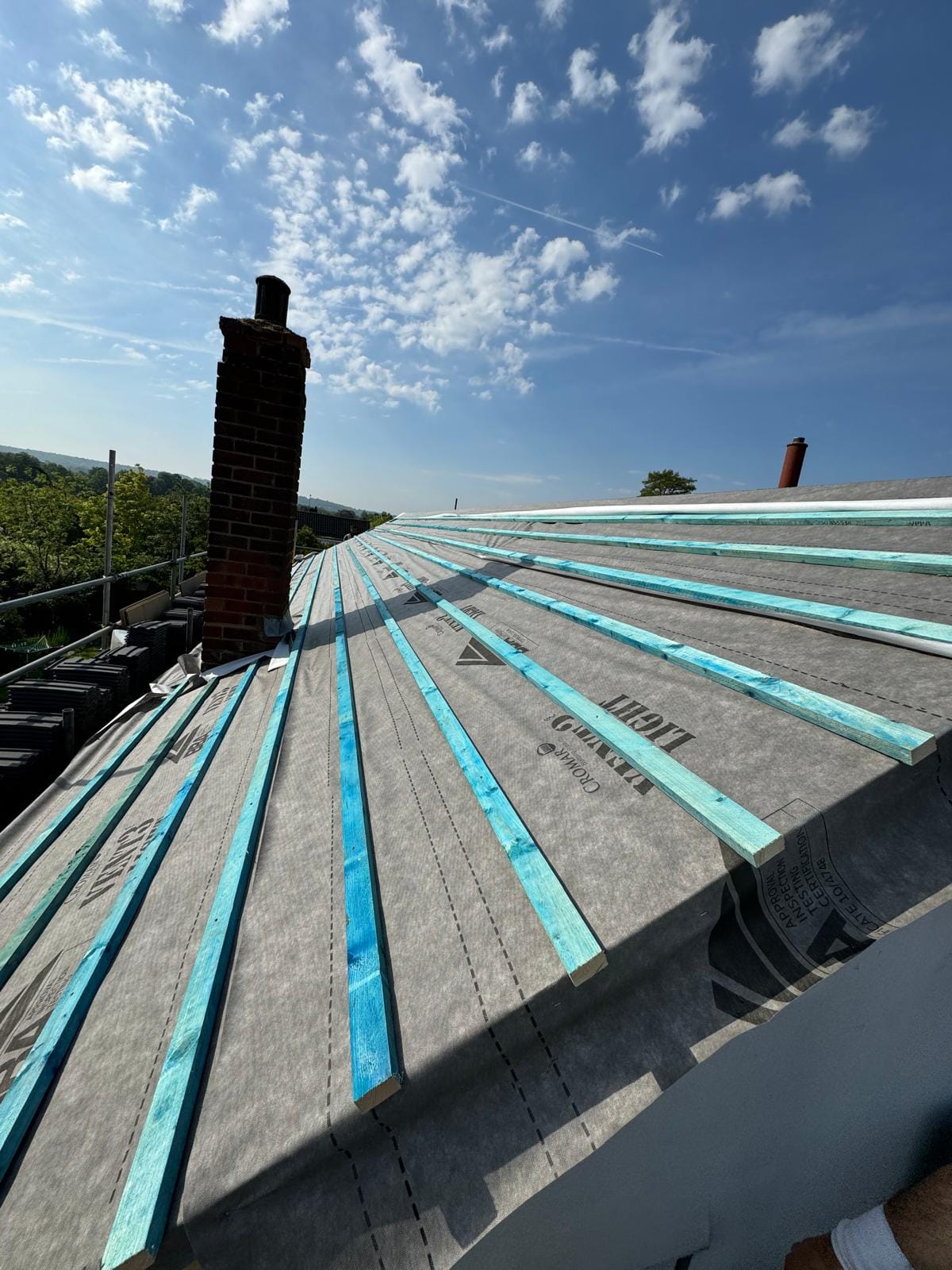 Pitched roof covered in grey breathable membrane with blue treated tile battens, beside a brick chimney stack.
