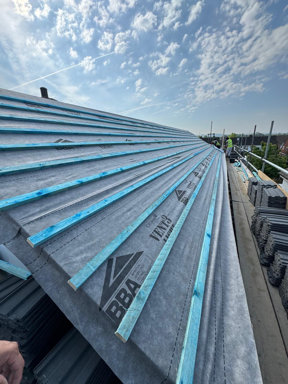 Roof slope in grey breathable membrane with blue tile battens, and concrete tiles stacked on the scaffold alongside.