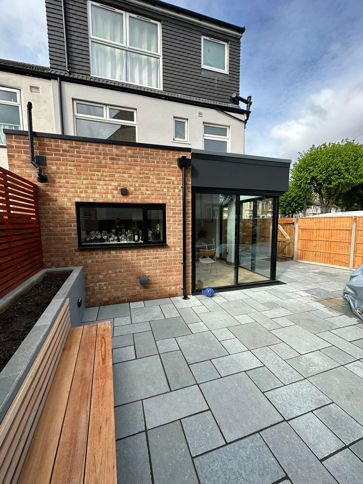 Single-storey red-brick rear extension with black-framed glazing and a dark fascia, opening onto a sandstone patio.