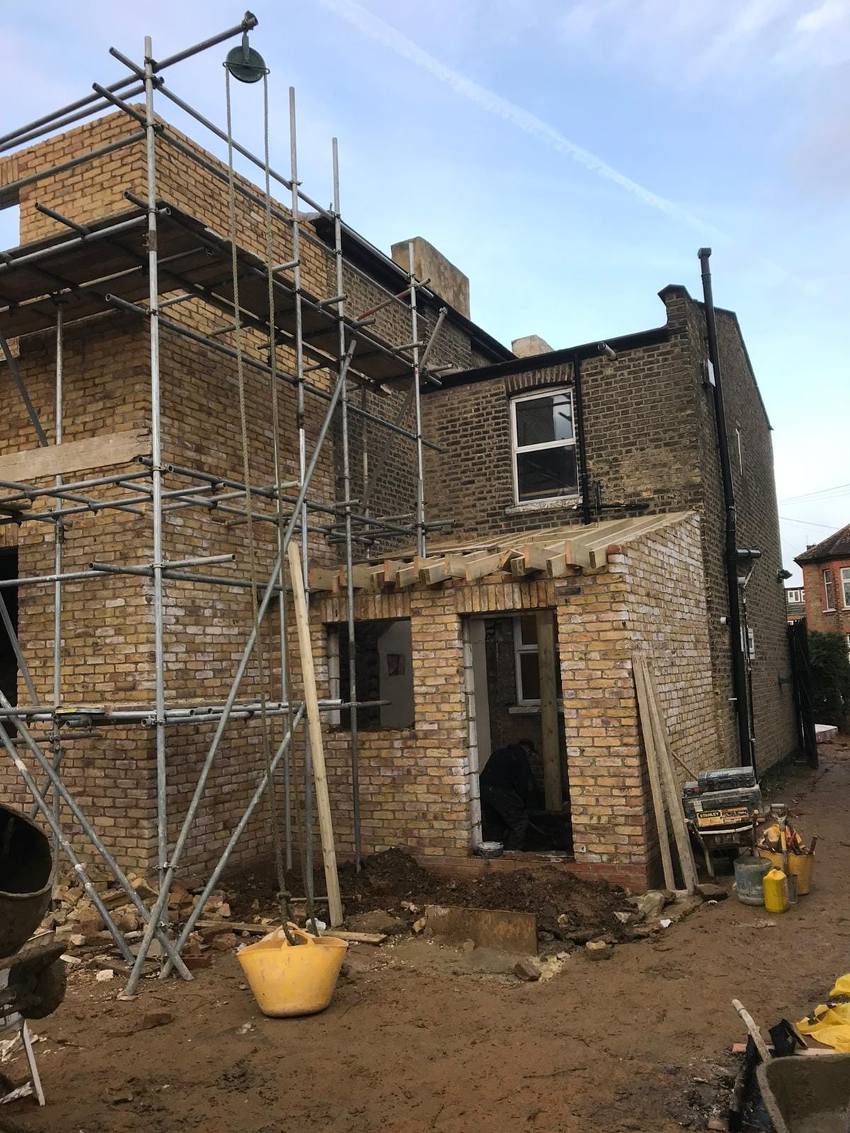 Stock brick two-storey structure under scaffold, with a single-storey section in front whose flat-roof joists are in place.