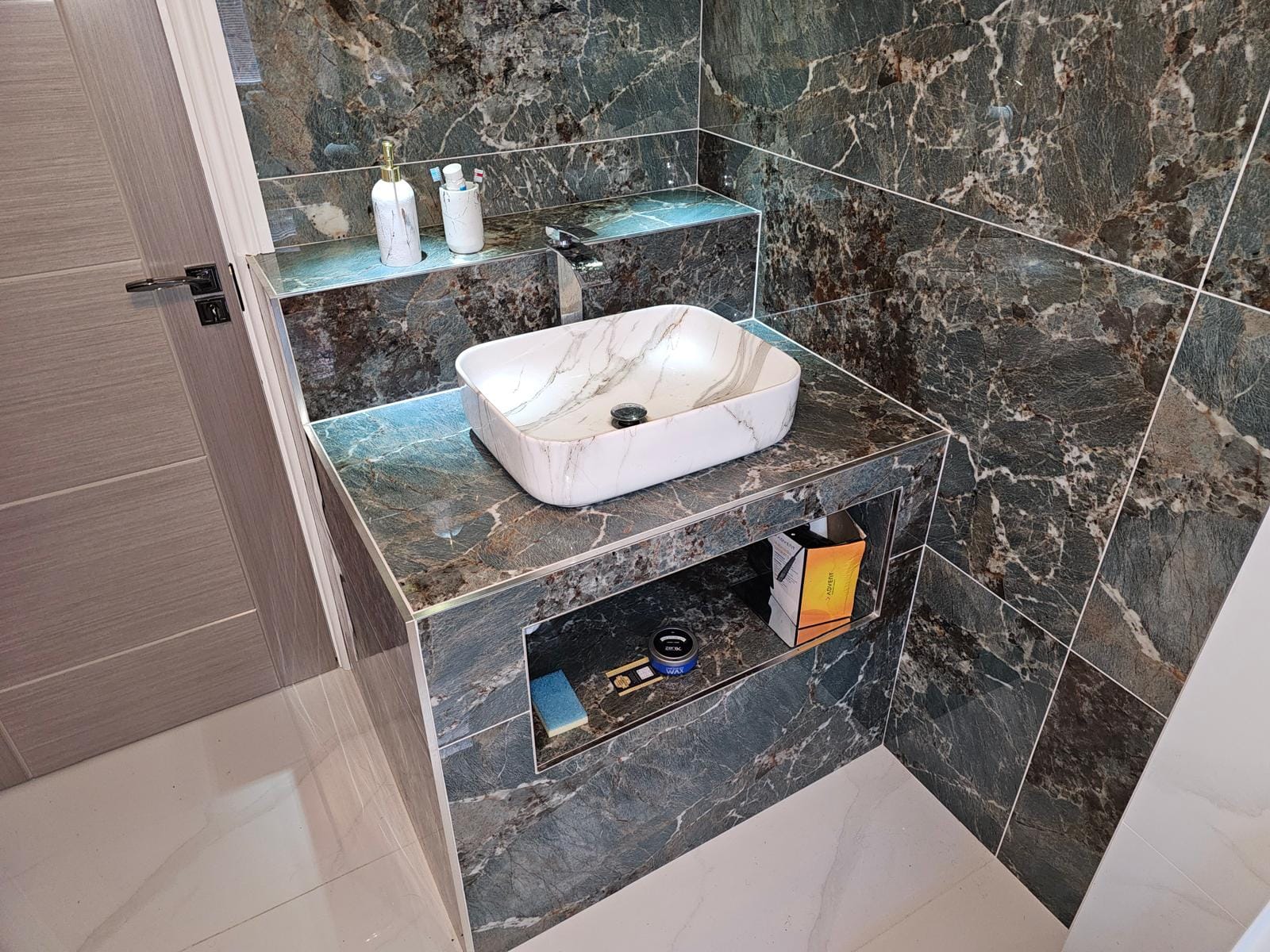 Tiled vanity plinth in dark-green marble effect with a vessel basin, lit ledge and open niche beside a grey door.