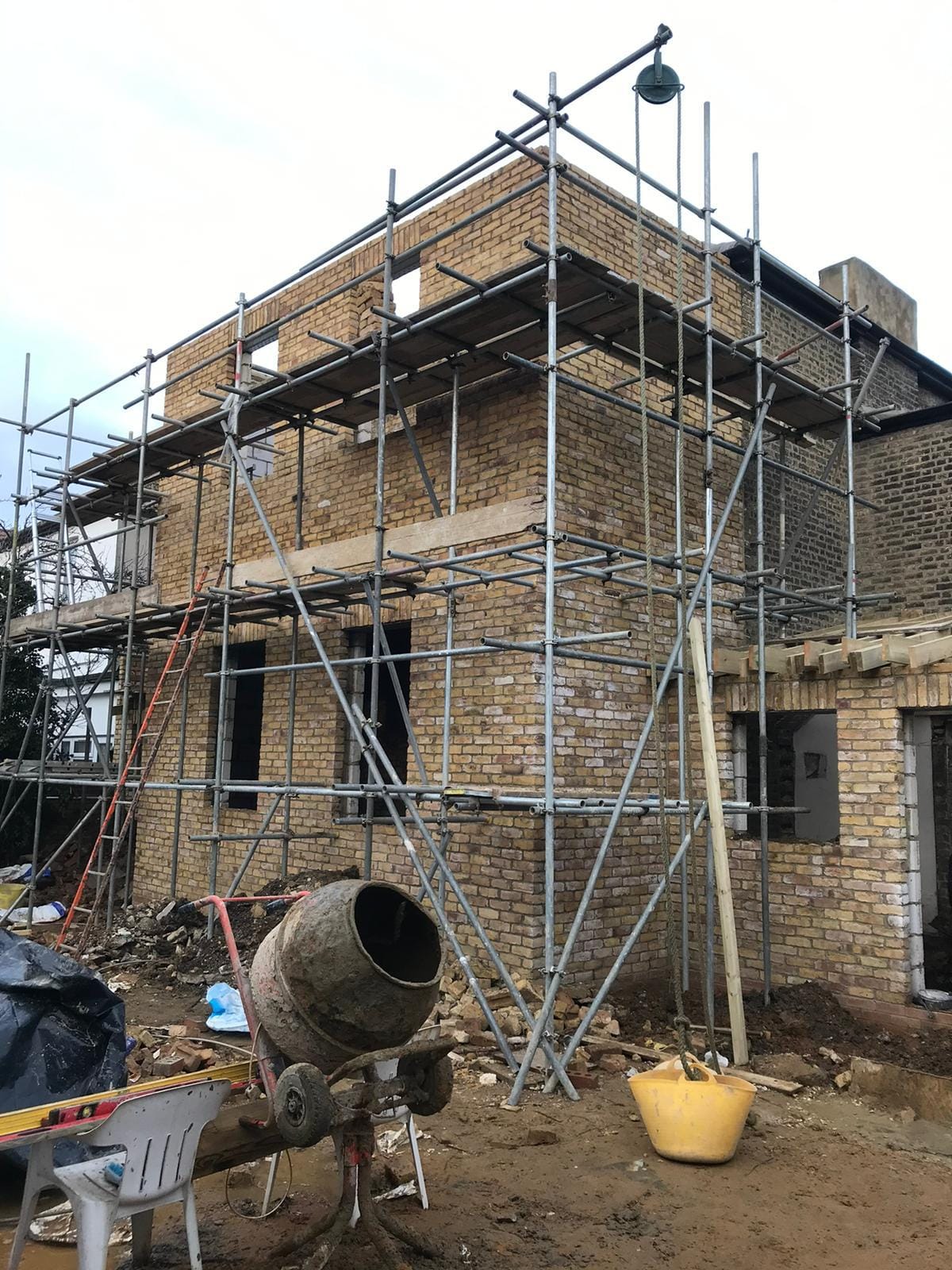 Two-storey structure in yellow stock brick under scaffold, with window openings formed and a cement mixer in front.
