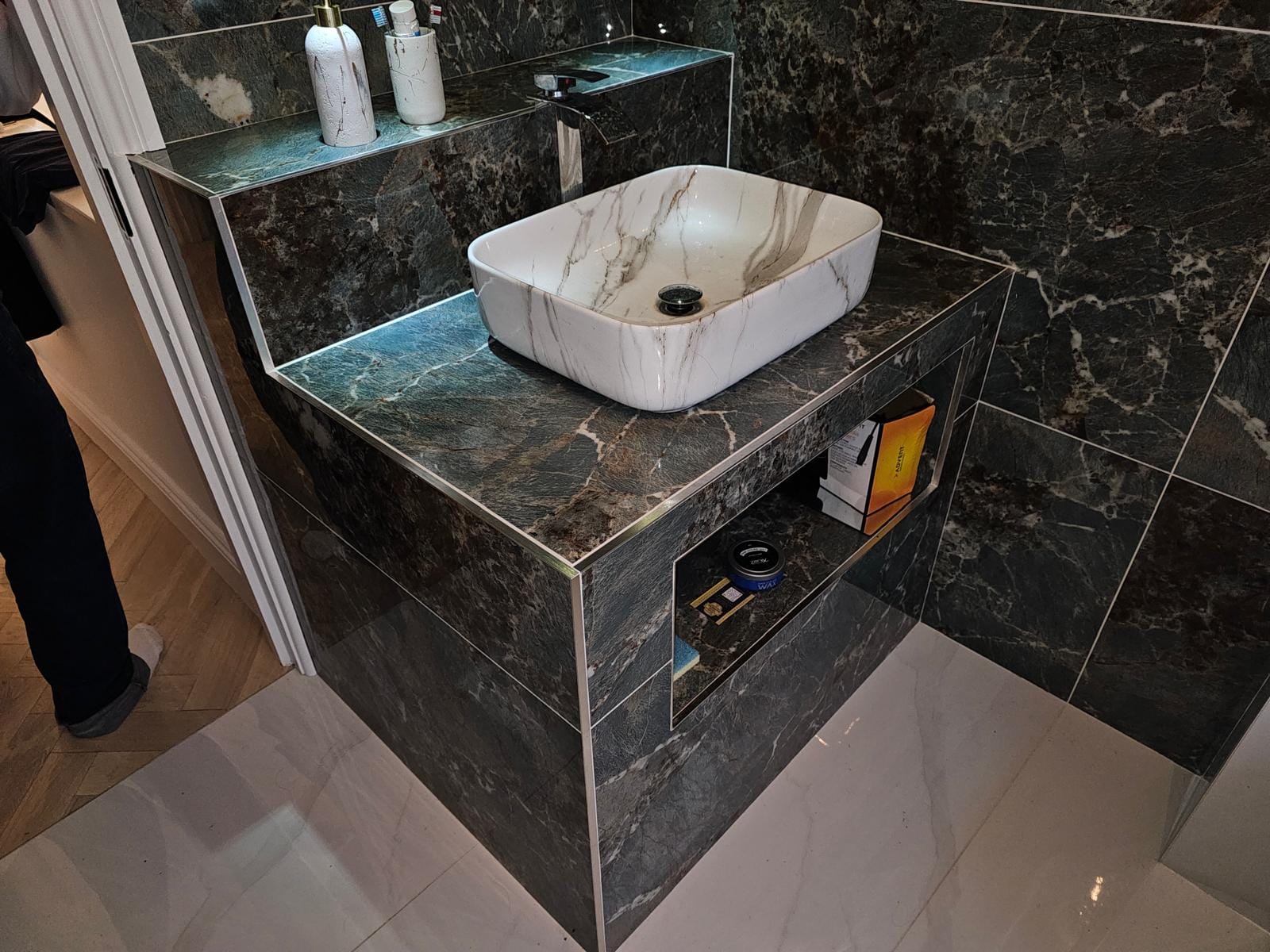 Close view of a marble-effect vessel basin on a dark-green tiled vanity plinth with a lit shelf and niche.