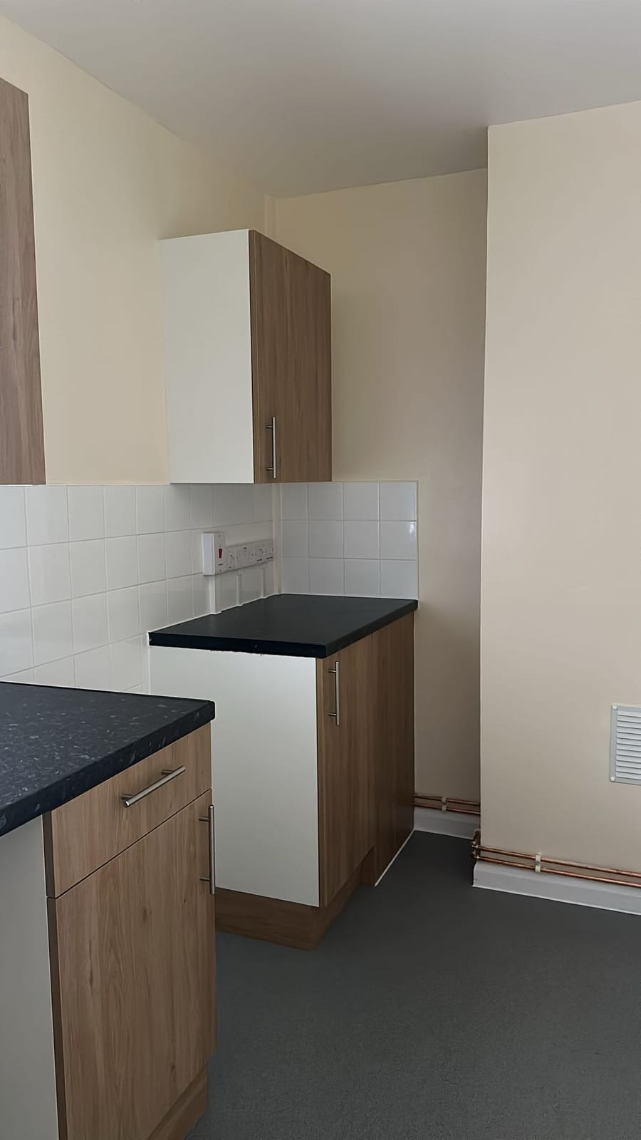 Refurbished kitchen in an empty flat with oak-effect units, a dark worktop, white tiles and grey safety flooring.